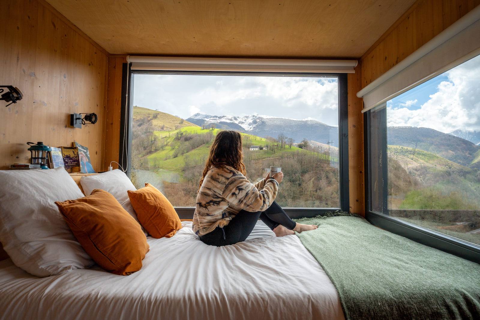 Una donna siede sul letto all'interno della minuscola casa, con una tazza in mano, con una vista mozzafiato sulle montagne.