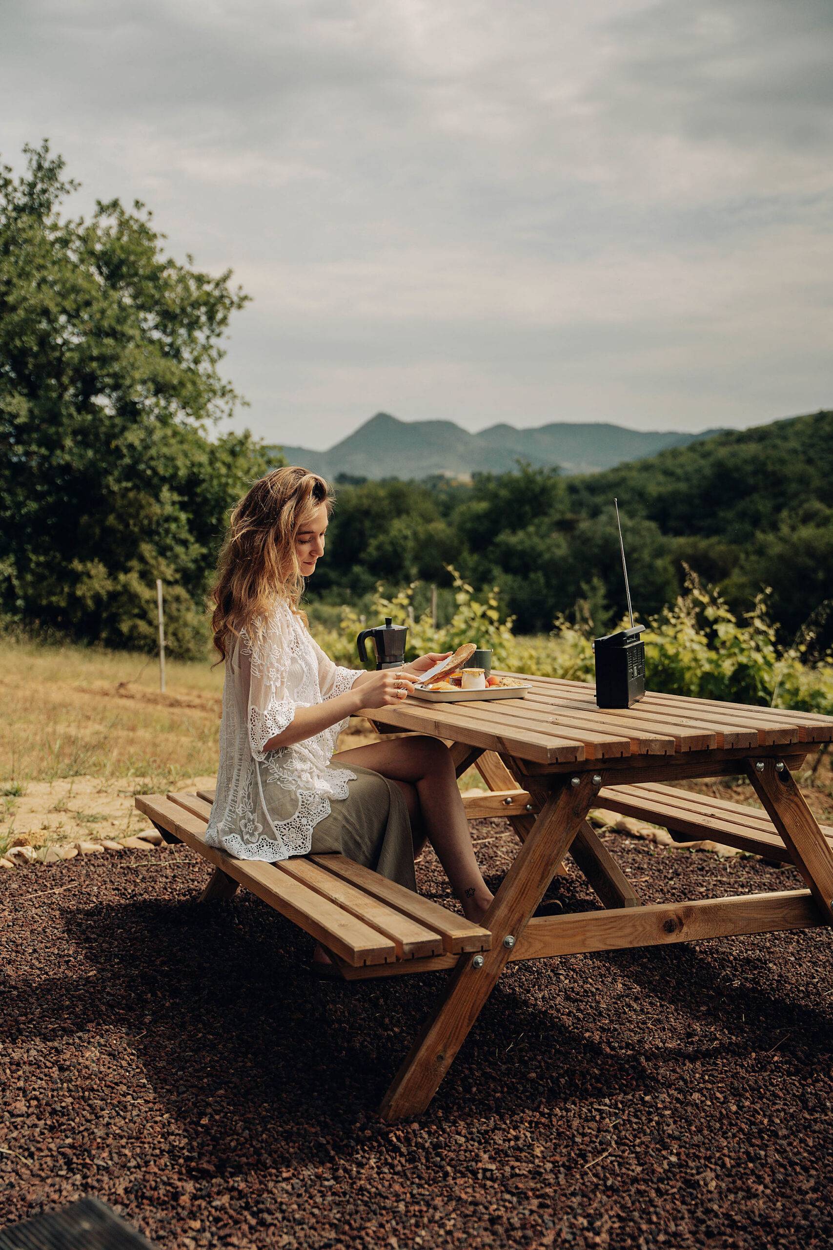 une femme est assise à une table en bois dehors, dans la nature, elle déjeune et écoute de la musique grâce au lecteur de k7, il y a la montagne en arrière plan