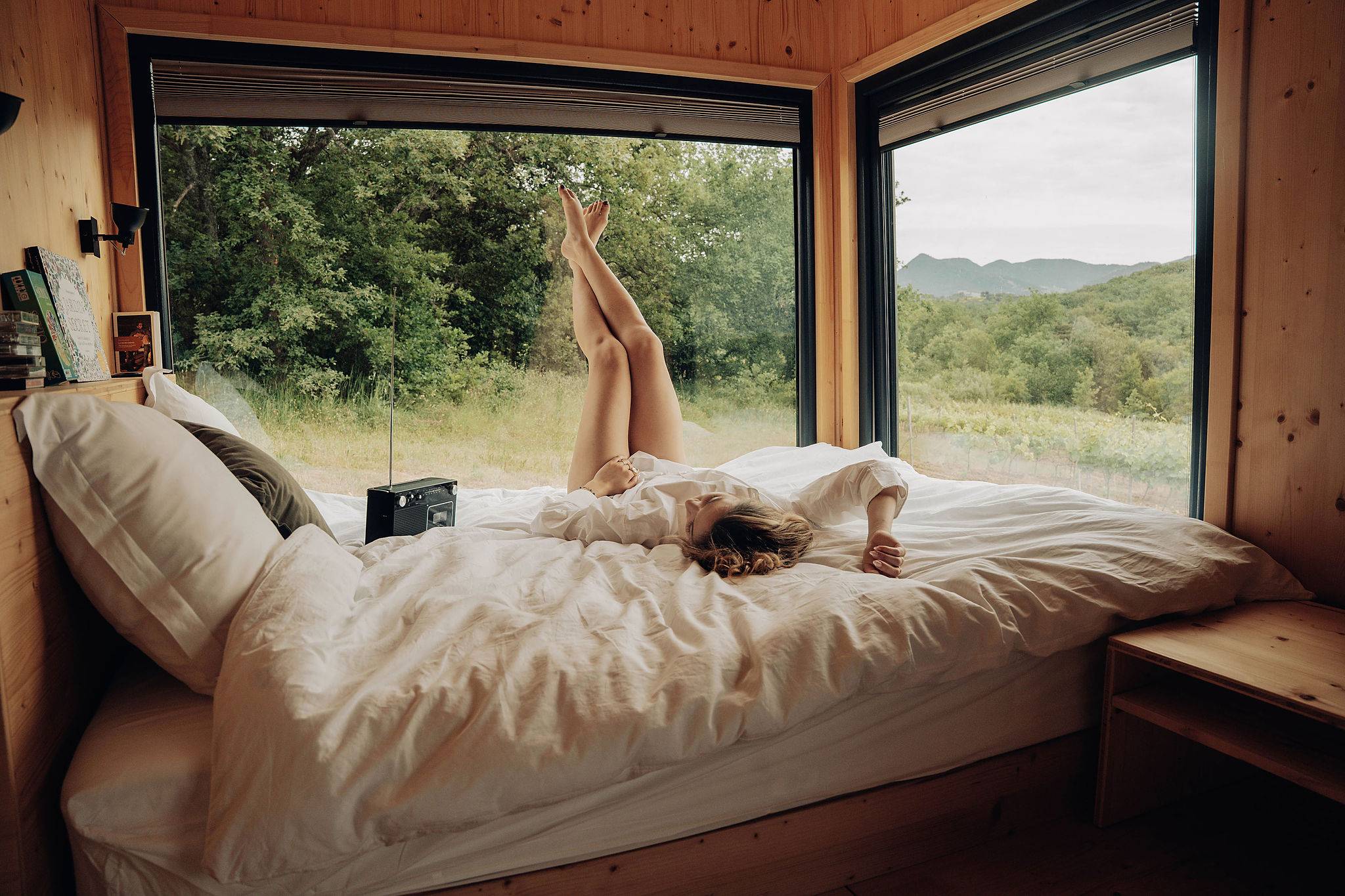 une femme est allongée sur le lit à l'intérieur de la tiny house, elle à les jambes croisés en l'air face aux fenêtre qui donnent vue sur la nature, elle écoute de la musique, le lecteur K7 est posé sur le lit à côté d'elle