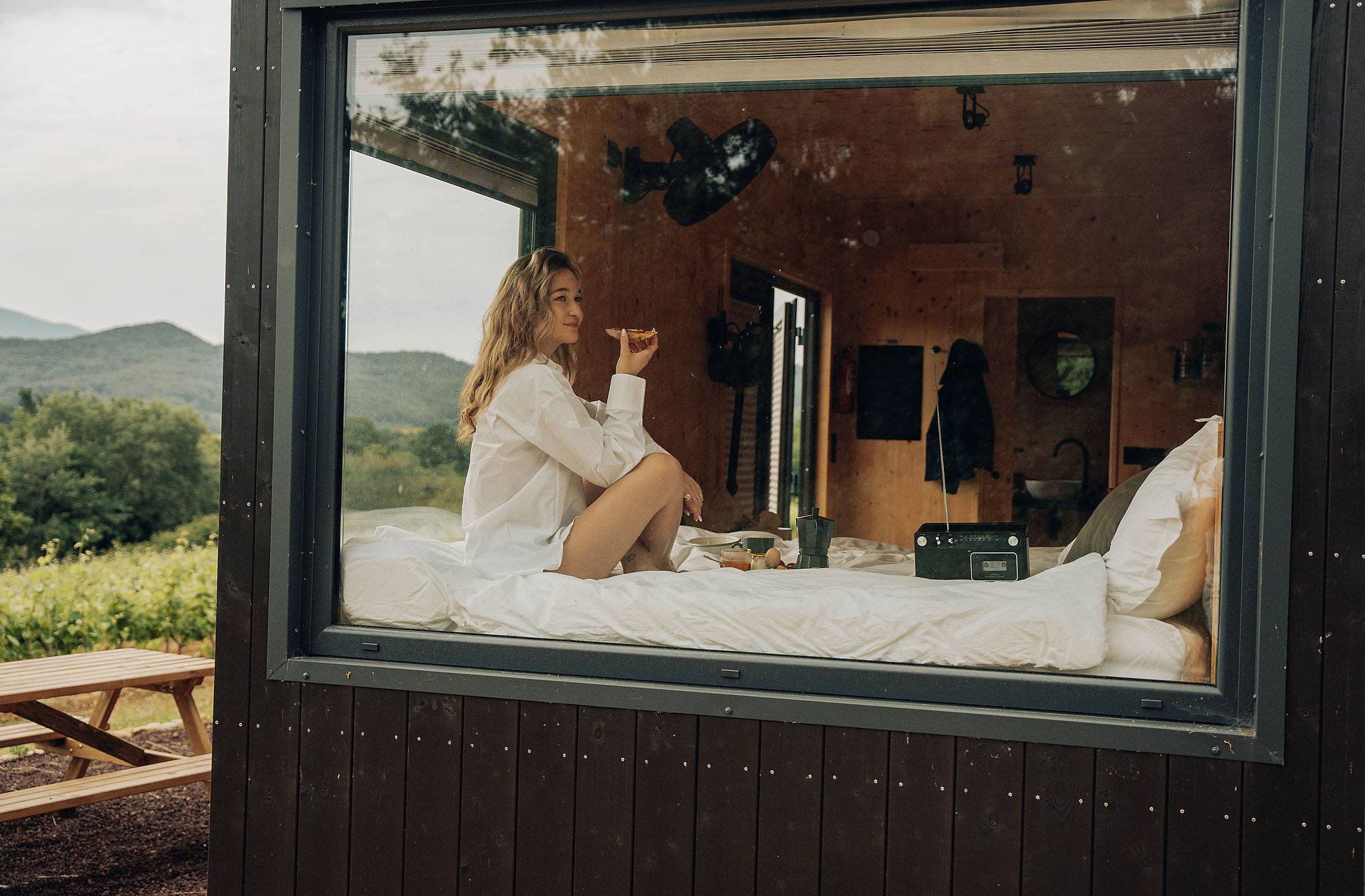 depuis l'extérieur on voit une femme assise sur le lit de la tiny house en train de manger son petit déjeuner et d'écouter la musique avec le lecteur de k7