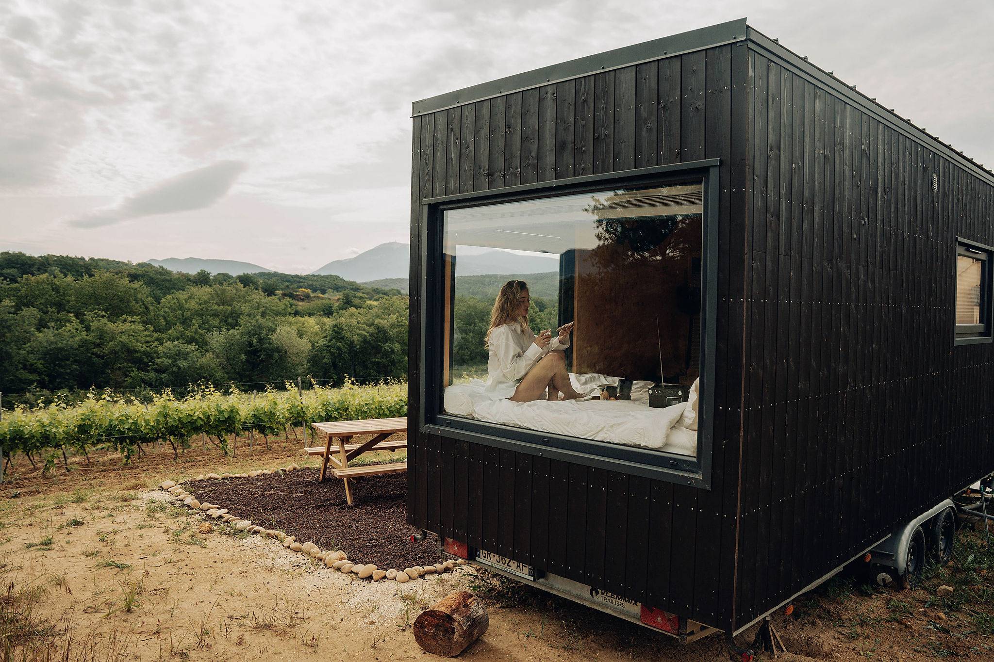 depuis l'extérieur on voit une femme assise sur le lit de la tiny house en train de manger son petit déjeuner et d'écouter la musique avec le lecteur de k7