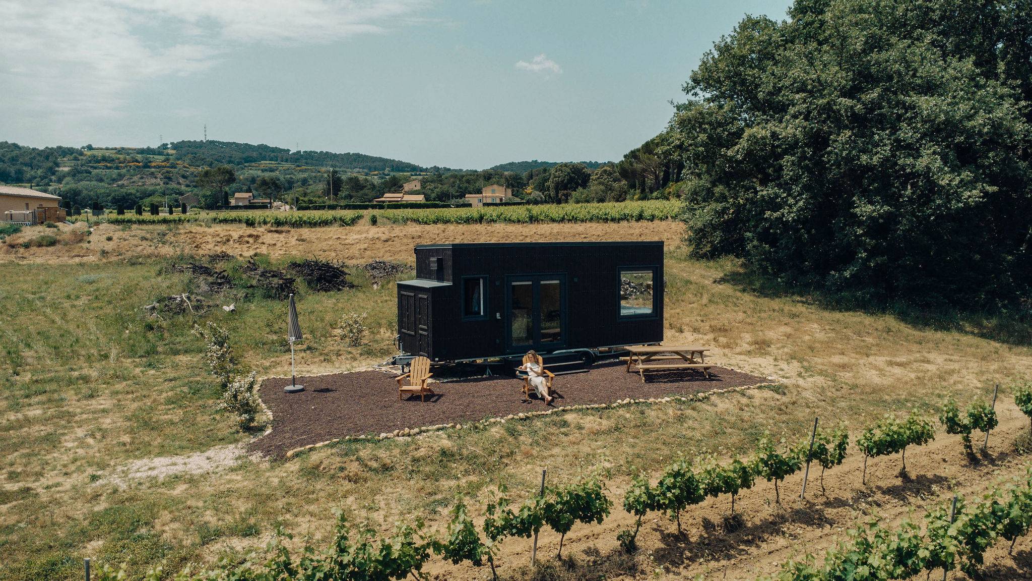vue de drone, il y a une tiny house un peu éloigné dans les vignes avec une femme assise sur une chaise en bois devant