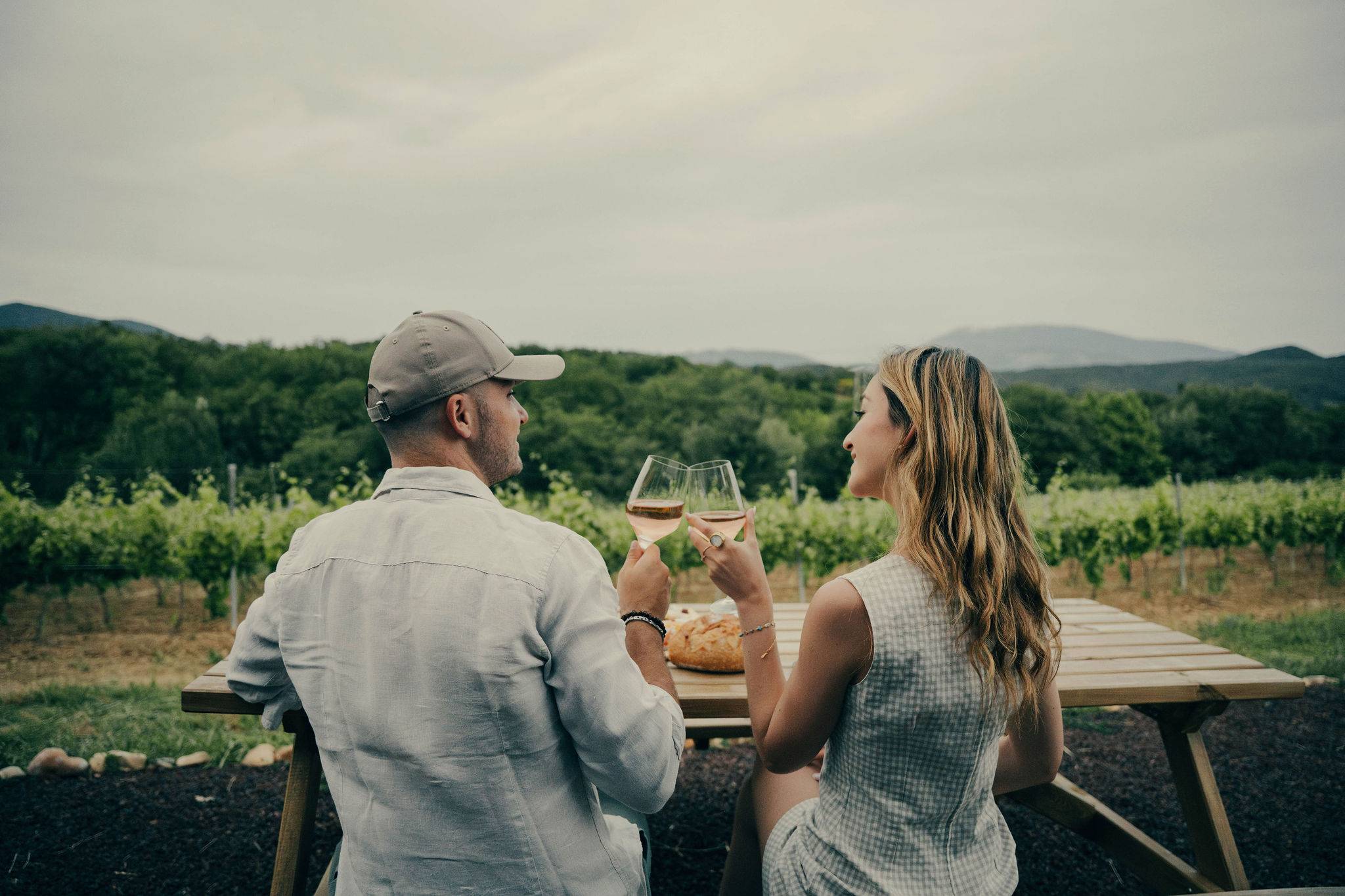 Un homme et une femme sont assis à une table en bois en extérieur, face à la montagne, ils trinquent avec un verre de vin à la main et ils se regardent