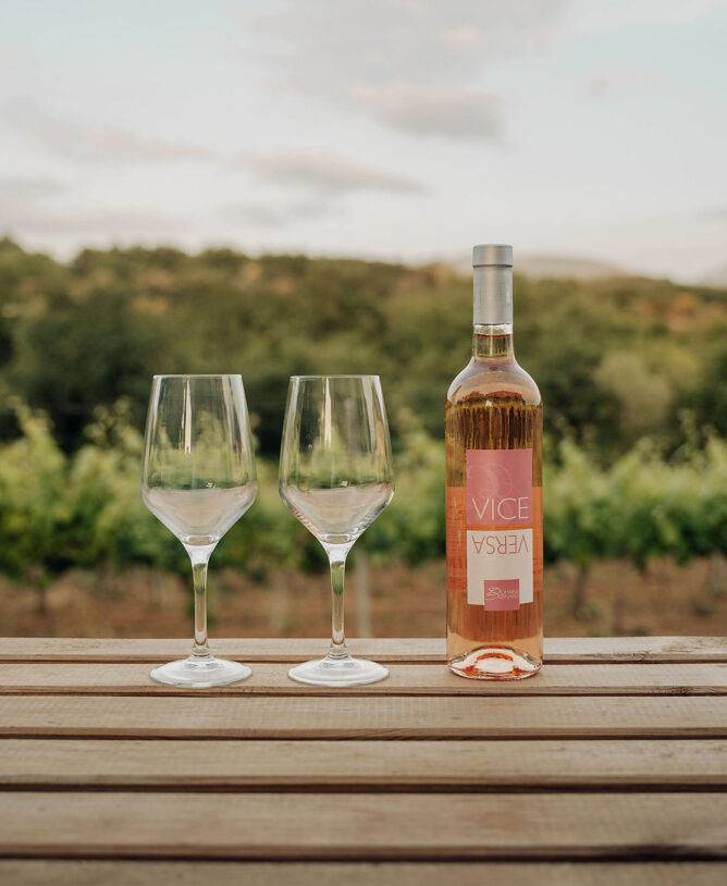 een houten tafel buiten, met een fles lokale rosé en twee glazen wijn, met uitzicht op de wijnranken en de bergen
